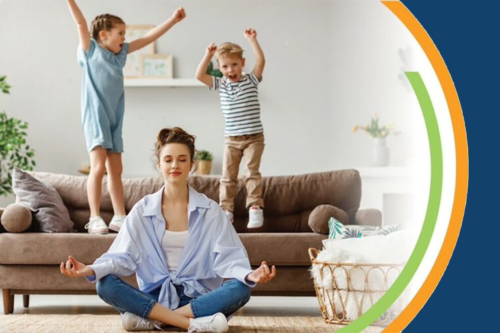 A mom sitting on the floor relaxing, while the children jump on the furniture in the background. 